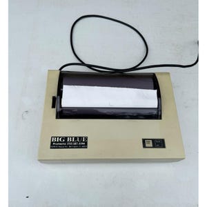 May include: A beige Big Blue Protecto printer with a black power cord. The printer has a black top with a roll of white paper visible. The Big Blue logo and contact information are on the front.