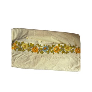 May include: A white fitted sheet with a floral border in shades of yellow, orange, and blue. The border features a repeating pattern of flowers and leaves.