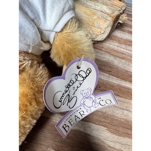 May include: A close-up of a plush teddy bear with light brown fur and a white shirt. A heart-shaped tag with cursive writing and a bear logo is attached. The tag reads "Collectible Bear Co."