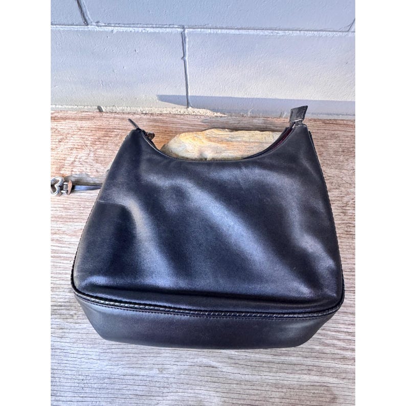 May include: Black leather shoulder bag with a curved top and a rectangular base. The bag has a single strap and a zipper closure. The bag is sitting on a wooden surface.