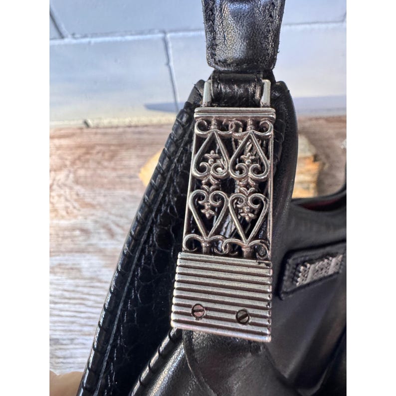 May include: Close-up of a black leather handbag with a decorative silver metal clasp. The clasp features an ornate design with heart-shaped elements and horizontal lines. The bag's leather has a textured appearance.