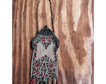 May include: A vintage metal mesh handbag featuring a floral pattern in red, green, and white. The bag has a dark metal frame with an intricate embossed design, a thin chain handle, and small red bead accents hanging from the bottom edge.