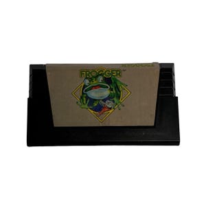 May include: A black video game cartridge with a brown label featuring the game title "FROOGER" and a cartoon frog graphic. The frog is green with a wide open mouth and is set against a yellow diamond.