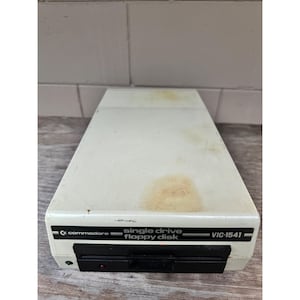May include: Off-white Commodore VIC-1541 single drive floppy disk. The drive has a black slot for inserting the disk and a red power indicator light. The Commodore logo and model information are printed on the front.