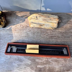 May include: A vintage slide rule in a brown case. The slide rule is black with white markings and a metal slider. The case is rectangular and has a reddish-brown exterior. A piece of light-colored paper is inside the case.