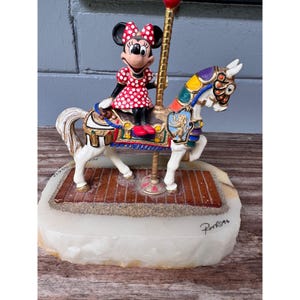 K&ouml;nnte beinhalten: Eine Minnie-Maus-Figur auf einem Karussellpferd. Das Pferd ist wei&szlig; mit bunten Akzenten und einem detaillierten Sattel. Minnie tr&auml;gt ein rotes Polka-Dot-Kleid und eine Schleife. Der Sockel ist ein hellfarbiger Stein mit einer ziegelartigen Plattform.