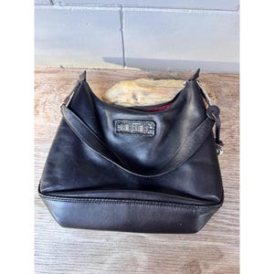 May include: Black leather shoulder bag with a rectangular emblem on the front. The bag has a curved shape, a single shoulder strap, and a red interior lining. The bag is sitting on a wooden surface.