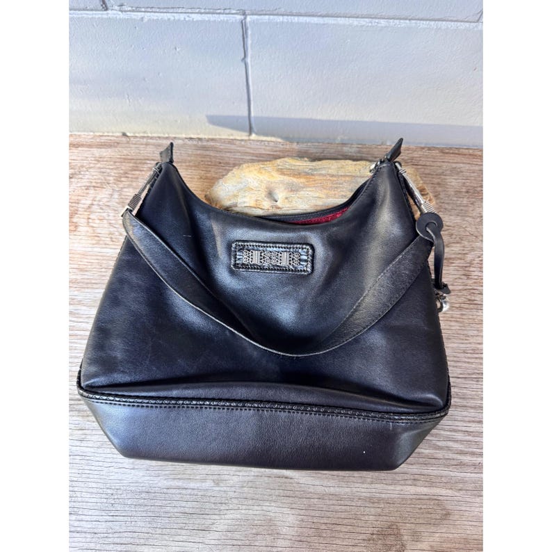 May include: Black leather shoulder bag with a rectangular emblem on the front. The bag has a curved shape, a single shoulder strap, and a red interior lining. The bag is sitting on a wooden surface.