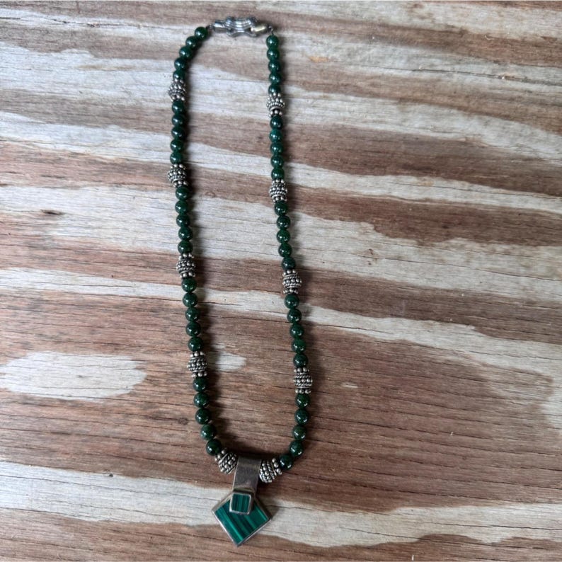 May include: A dark green beaded necklace with silver-colored accents and a square pendant. The pendant features a green stone inlay. The necklace is displayed on a wooden surface.