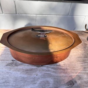 May include: Oval copper cooking pot with a matching lid. The pot has a dark handle and a shiny copper finish. The pot is sitting on a wooden surface. The pot is likely used for cooking.