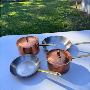 May include: Four pieces of cookware, including two copper pots with lids and two stainless steel pans with brass handles, are arranged on a white surface. The copper pots have a warm, reddish-brown hue, while the pans have a silver color.
