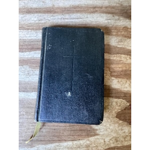 May include: A small, black book with a cross embossed on the cover. The book has a gold ribbon bookmark and shows signs of wear. The cover is slightly textured and the edges are worn.