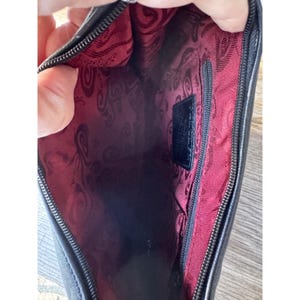 May include: Black leather clutch with a red patterned interior. The interior features a zippered pocket and a small black label. The clutch is open, revealing the interior lining and the zipper.