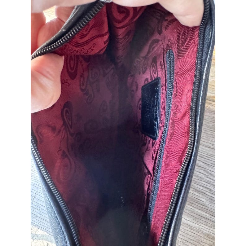 May include: Black leather clutch with a red patterned interior. The interior features a zippered pocket and a small black label. The clutch is open, revealing the interior lining and the zipper.
