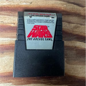 May include: A black Atari 2600 game cartridge with a white label that reads "STAR WARS THE ARCADE GAME". The label has a red "STAR WARS" logo.