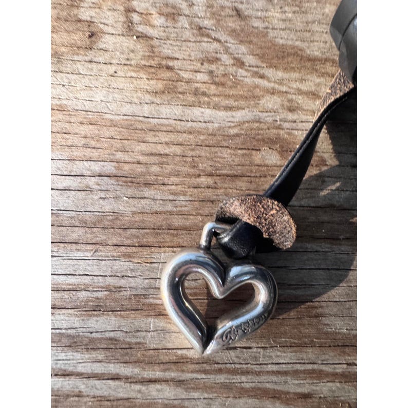 May include: A silver heart pendant with a black leather strap. The heart-shaped charm has a cutout center and is attached to a black leather strap with a brown leather accent. The pendant is on a wooden surface.
