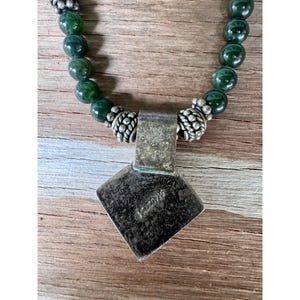May include: A necklace featuring a pendant with a square, textured silver pendant and a string of green beads. The pendant is attached to the bead strand with silver connectors. The necklace rests on a wooden surface.