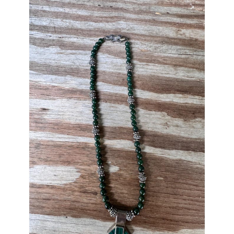 May include: A green beaded necklace with silver accents and a geometric pendant. The necklace features small, round green beads interspersed with silver-colored decorative beads. The pendant is a silver-toned metal with a green stone.