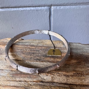 May include: Silver-toned bracelet with a textured, bark-like design. The bracelet has a series of raised, rectangular accents. A small tag with a black string is attached. The bracelet is displayed on a piece of wood.