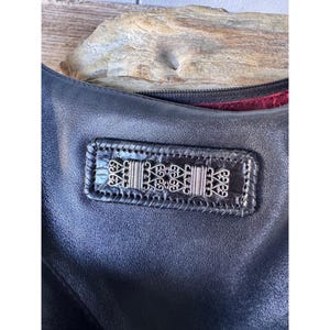 May include: Black leather handbag with a decorative silver metal accent. The bag features a rectangular patch with intricate silver filigree designs. The leather has a subtle texture, and the bag's interior is lined with a red fabric.