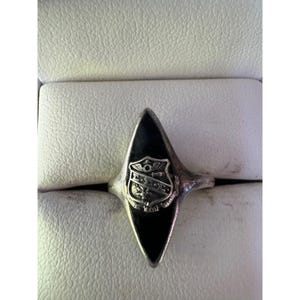 May include: Silver-toned ring with a black, elongated, diamond-shaped face. A shield emblem is centered on the face. The ring is set against a white, textured background.