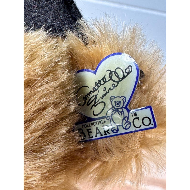 May include: Close-up of a plush teddy bear with tan fur and a black cap. A heart-shaped tag with the text "Symette Zuniga" and a bear logo is attached to the bear. The tag also reads "Collectible Bears Co."