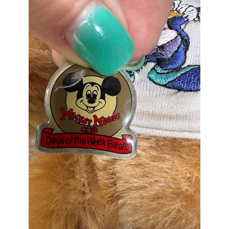 May include: A close-up of a plush bear with a Mickey Mouse Club tag. The tag is gold with a Mickey Mouse graphic and the words "Mickey Mouse Club" and "Days of the Week Bears" in red. The bear is light brown.