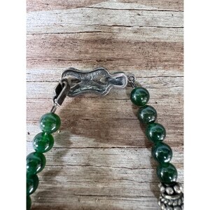 May include: A close-up of a necklace featuring large, round, dark green beads. The necklace has a silver-colored clasp and decorative elements. The beads are strung together, creating a bold and eye-catching piece of jewelry.