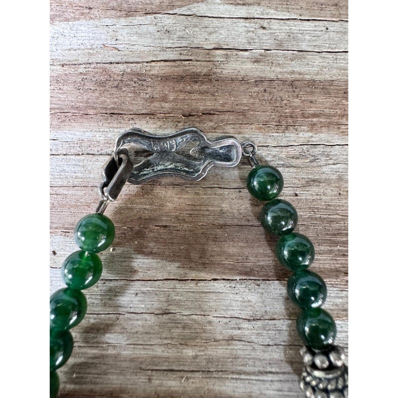 May include: A close-up of a necklace featuring large, round, dark green beads. The necklace has a silver-colored clasp and decorative elements. The beads are strung together, creating a bold and eye-catching piece of jewelry.