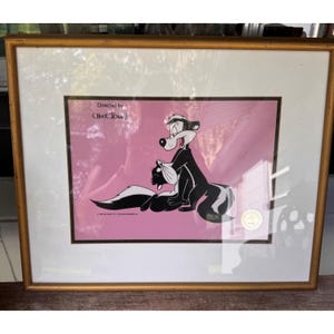 May include: A framed animation cel featuring Pepe Le Pew holding a smaller skunk against a solid pink background. The text Directed by Chuck Jones is printed in black in the upper left corner. The artwork is enclosed in a gold-toned frame.