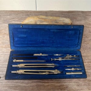 May include: A vintage drafting set in a blue velvet-lined case. The set includes several compasses, dividers, and other drawing tools. The case is open, revealing the tools neatly arranged. The tools are made of metal and have a silver and black finish.