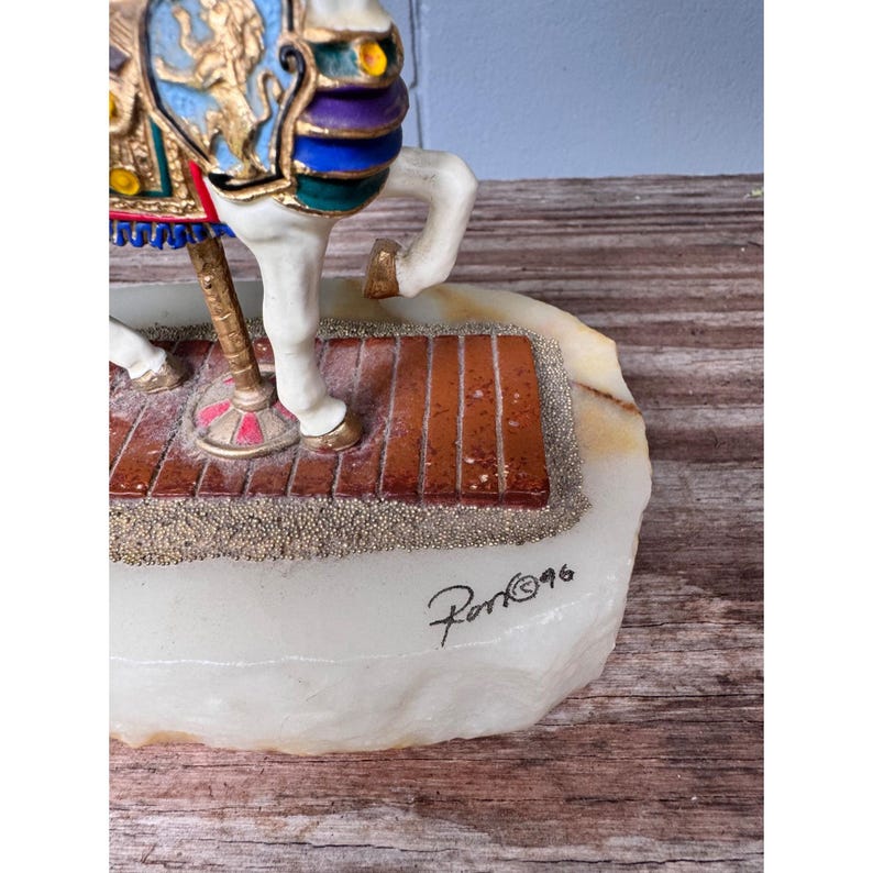 K&ouml;nnte beinhalten: Dekorative Karussellpferde-Figur mit wei&szlig;em K&ouml;rper und farbenfrohen Akzenten. Das Pferd hat eine goldfarbene Stange und steht auf einem ziegelgemusterten Sockel. Der Sockel steht auf einem wei&szlig;en Stein mit der Signatur "Ron&copy;96".