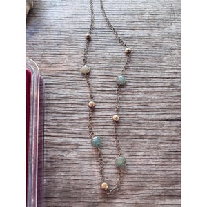 May include: A long necklace with a delicate chain, featuring alternating gold-colored beads and round, pale green gemstones. The necklace is displayed on a wooden surface, showcasing its elegant design and color contrast.