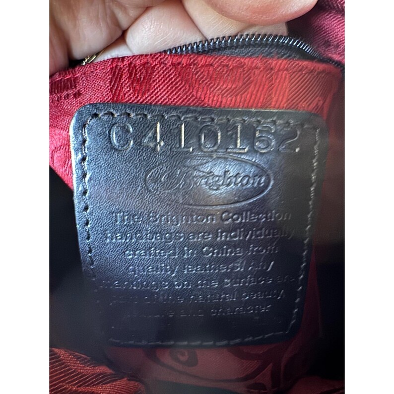 May include: Close-up of a black leather tag with the text "C410152" and the Brighton logo. The tag is stitched onto a red fabric with a floral pattern. The text "The Brighton Collection" is also visible.