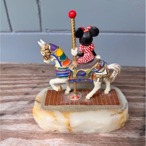 K&ouml;nnte beinhalten: Ein Karussell-Figur mit Minnie Mouse, die auf einem wei&szlig;en Pferd mit bunten Akzenten reitet. Das Pferd hat einen dekorativen Sattel und ist auf einem Sockel mit Ziegelmuster montiert. Die Figur tr&auml;gt ein rot-wei&szlig; gepunktetes Kleid.