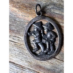 May include: Oval silver pendant featuring two figures in hats, one playing a horn and the other a drum. The pendant has a loop at the top for a chain. The background is a weathered wood surface.