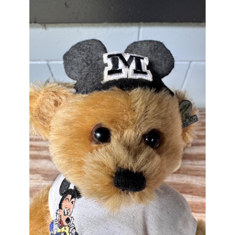 May include: A plush teddy bear with brown fur, wearing a gray hat with a white "M" and a white t-shirt with a Mickey Mouse graphic. The bear has large black eyes and a black nose.