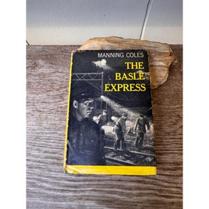 May include: A hardcover book titled The Basle Express by Manning Coles. The black and white cover features a dramatic illustration of three figures in uniform on train tracks at night, illuminated by a bright spotlight. The title is in yellow text.