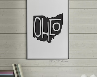 Puede incluir: Lámina artística enmarcada en blanco y negro con el contorno del estado de Ohio y la palabra "OHIO" en una fuente audaz y estilizada. La impresión se muestra en una pared blanca con un fondo de paneles de madera. Las dimensiones son 61 cm x 91 cm.