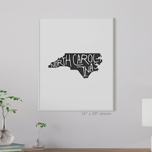 May include: A framed print showing the silhouette of North Carolina in black, with the state's name written inside. The print is set against a white backdrop, with dimensions of 40.6 x 50.8 cm shown. The artwork is a minimalist design.