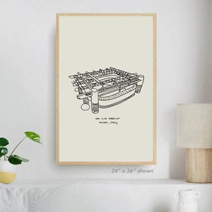 Könnte beinhalten: Ein gerahmter Kunstdruck mit einer Schwarz-Weiß-Strichzeichnung des San Siro Stadions in Mailand, Italien. Das Kunstwerk wird auf einem cremefarbenen Hintergrund angezeigt und ist mit "24" x 36" shown" beschriftet. Die Stadionillustration ist detailliert und architektonisch.