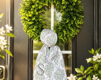 May include: A vibrant green wreath made of artificial leaves hangs on a black door. A decorative white and green patterned fabric with the letter "S" is attached to the wreath. White flowers and greenery are visible on the sides.