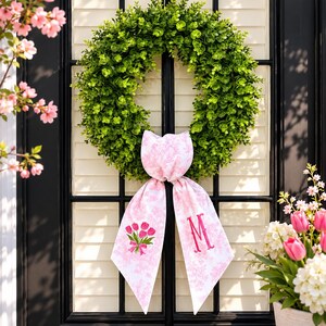 May include: A green, leafy wreath with a pink and white patterned bow, adorned with a bouquet of tulips and the letter "M". The wreath hangs on a black door with white shutters, with pink flowers in the background.