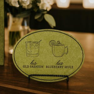 May include: A green oval sign with illustrations of cocktails, labeled "his" and "her" with "Old Fashion" and "Blueberry Mule" text. The sign is on a black stand, with a floral arrangement and illuminated "LOVE" letters in the background.