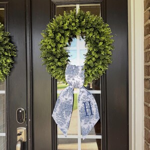 May include: A green wreath with a white and blue patterned bow, featuring the letter "H" in the center, hangs on a black door. The wreath is made of small, round green leaves. The bow has a toile pattern.