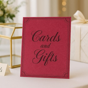 May include: A burgundy sign with the words "Cards and Gifts" in elegant script. A small card with "Cards" is in front of the sign. A gold geometric terrarium and a wrapped gift box with a cream ribbon are also present.
