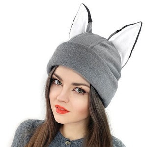 May include: A gray fleece beanie with two white and black furry ears. The beanie is worn by a person with long brown hair.