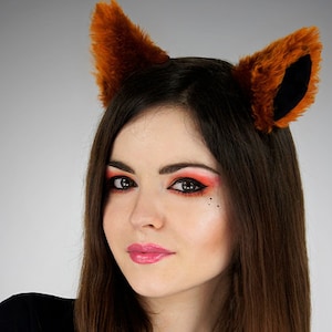 Op de afbeelding: Een vrouw met lang bruin haar die een zwart shirt en vossenoren-hoofdbanden draagt. Ze heeft rood en oranje oogschaduw en zwarte eyeliner.