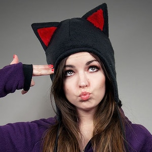 May include: Black cat ear hat with red inner ears. The hat is made of a soft material and has a hood. The person in the image is wearing a purple fleece jacket.