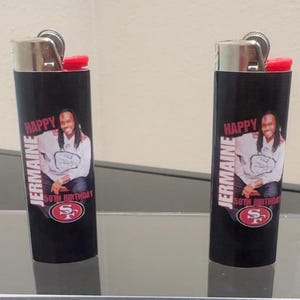 May include: Two black lighters with red tops. Each lighter has a personalized label with a photo of a man, the text "Jermaine", "Happy 50th Birthday", and a red and white logo.